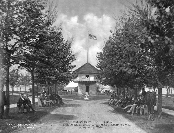 Front view of the Block House, the Pennsylvania Soldiers' and Sailors' Home. Men sit on benches along both sides of a path leading to the house, with a flag on the roof waving on a pole. Caption reads: "Block House. PA. Soldiers' and Sailors' Home. Erie, PA."