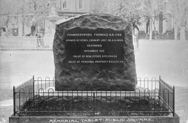 View of a memorial tablet commemorating the burning of the city by the Confederates. The memorial stands in a public square, surrounded by a fence. Behind the tablet are a fountain, trees, and buildings. Caption reads: "Memorial Tablet • Public Square." Tablet reads: "Chambersburg Founded A.D. 1764. Burned by Rebel Cavalry July 30, A.D. 1864. Destroyed Buildings 537. Value of Real Estate $713,294.34. Value of Personal Property $915,137.24."