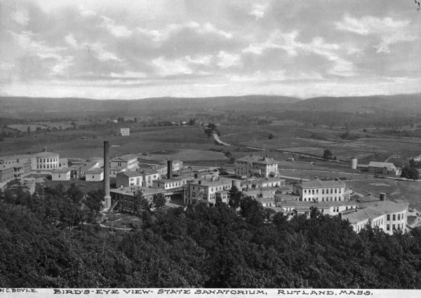 Elevated view of the State Sanatorium, constructed in 1898, and a hilly countryside. Below the trees in the foreground stand the many buildings of the sanatorium, as well as farm buildings and land. Caption reads, "Bird's-eye View - State Sanitorium, Rutland, Mass."