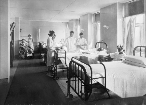 Interior of the largest ward in St. John's Hospital, started in 1952. Nurses and doctors are standing by patients. One of the patients is sitting next to a hospital bed, and another is lying in a bed. In the background a woman is standing near another person sitting in a wheelchair.