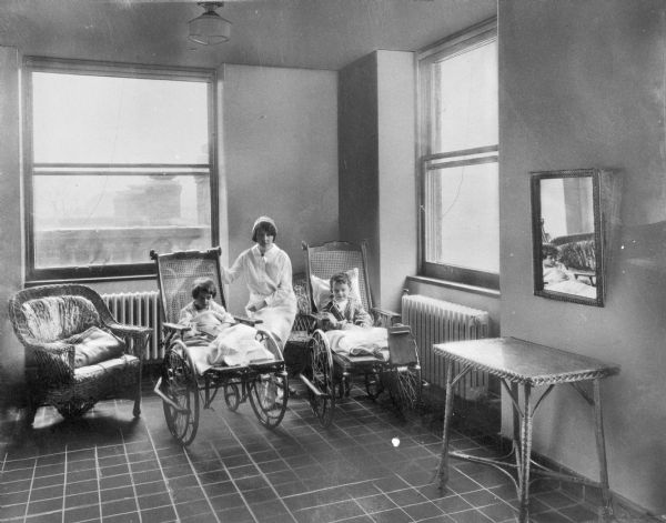 Interior of the sun parlor of the childrens' ward at St. John's Hospital, started in 1952. A nurse sits between two children in wheelchairs.