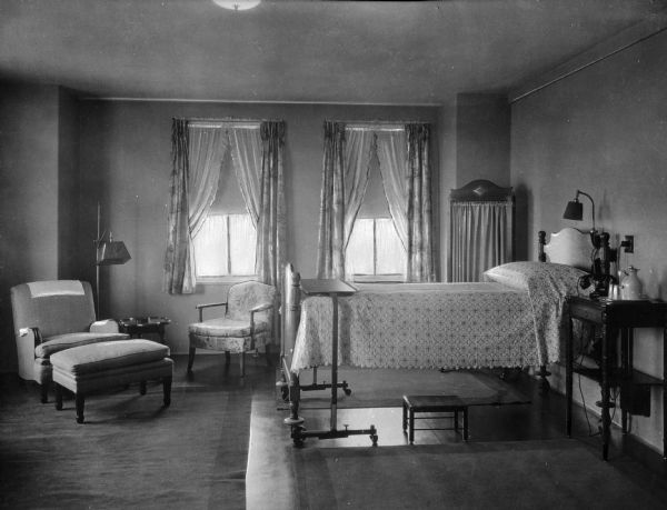 Interior of a bedroom at Doctor's Hospital, founded by Theodore Atlas in the early 1900's.  A view from across the room shows a patient's bed, two chairs, and windows.