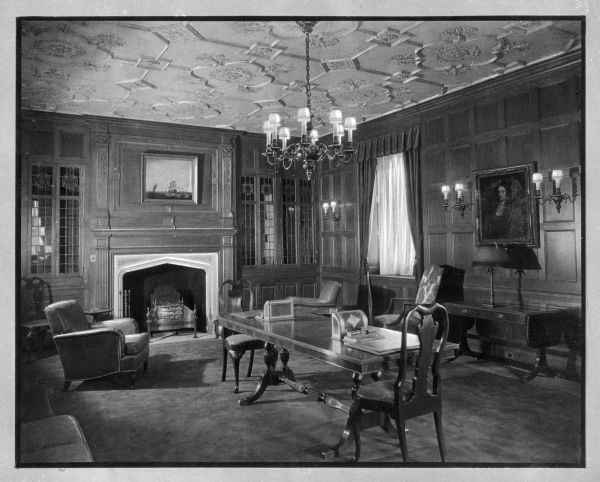 Interior of the library at the Doctor's Hospital, founded by Theodore Atlas in the early 1900's.  The view includes a table and chairs with a fireplace and window behind them.  A chandelier hangs from a decorative ceiling, and paintings are framed on the walls.