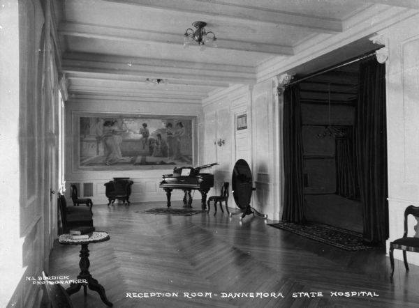 Interior of the reception room of Dannemora State Hospital, built in 1899 as a mental health facility. The view includes a large painting on the far wall near a piano, and chairs and tables are around the room. On the right, a set of open curtains frame the doorway to another room. Caption reads: "Reception Room — Dannemora State Hospital."