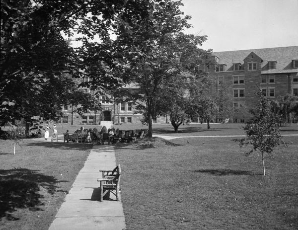 Slightly elevated view of the Philadelphia Home for Incurables, established in 1877. Nurses stand near a  group of patients sitting outdoors under trees, with some sitting in wheelchairs.