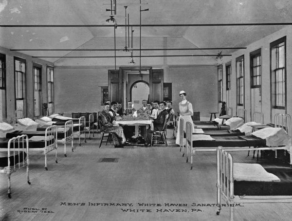 Interior of the men's infirmary at White Haven Sanatorium, established in 1901. A group of men sit at a table and a nurse stands beside them. Patient beds and windows line both sides of the room. Caption reads: "Men's Infirmary, White Haven Sanatorium, White Haven, PA."