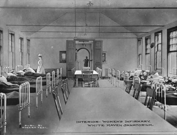 Interior of the women's infirmary at White Haven Sanatorium, established in 1901. The view includes patients in beds lining both sides of the room, a nurse, and a man standing in the doorway in the background. Caption reads: "Interior — Women's Infirmary, White Haven Sanatorium."