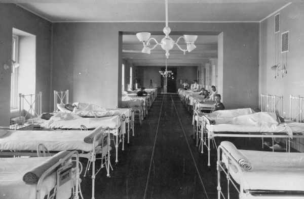 Interior of the general ward at the United States Naval Hospital, established in 1834 on the original site of Fort Nelson. Patients sit in chairs or lie in beds that line the walls on both sides of the room. The long walkway between beds leads to a door at the end of the ward.
