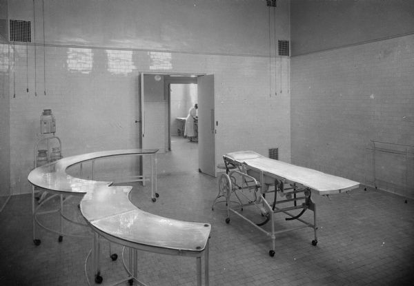 Interior of an operating room at the United States Naval Hospital, established in 1834 on the original site of Fort Nelson. The view includes operating tables, and through the doorway, a nurse standing next to a table.