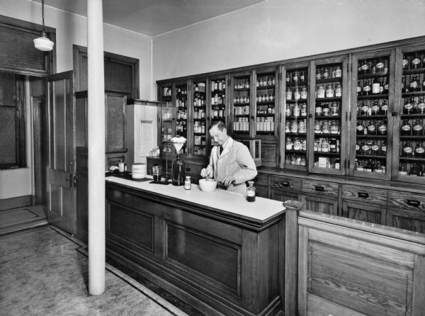 Interior of the pharmacy at Saint Mary's Hospital, completed May 18, 1859 and designed by Anthony Bley. A pharmacist works behind the counter. Behind him are cabinets along the wall holding medicine.