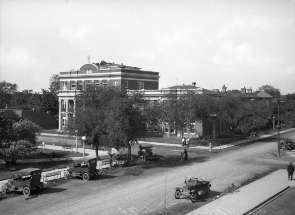 Elevated view of Mercy Hospital, founded by the Sisters of Mercy in 1894. An automobile turns the corner onto a street where cars are parked. Individuals cross the street and walk on the sidewalks.