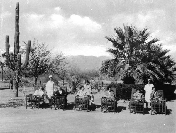 View of patients resting in chairs and nurses standing near them at Saint Mary's Sanatorium, founded in 1880 by Bishop Salpointe. A large cactus and a palm tree are in the background.