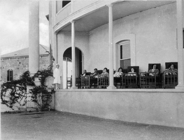 View from grounds toward patients sitting on the porch of the rotunda at Saint Mary's Sanatorium, founded in 1880 by Bishop Salpointe. A nurse stands near a column beside the relaxing patients.