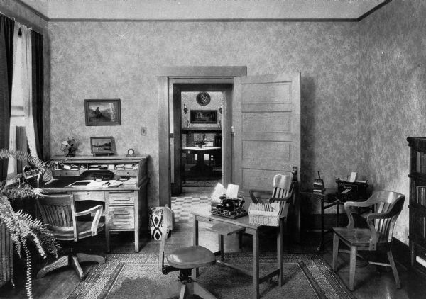 Interior view of an office at Saint Mary's Sanatorium, founded in 1880 by Bishop Salpointe. Near the window on the left is a desk, in the center is a typewriter on a table, and chairs and a bookcase are on the right. In the back right corner is a Burroughs Adding Machine. In the background an opened door leads to another room with a table.