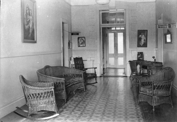 Interior of the waiting room at Saint Mary's Sanatorium, founded in 1880 by Bishop Salpointe. The room has wicker furniture and a wooden rocking chair and is decorated with religious paintings. A sign on the right reads "Pharmacy," and a sign on the left reads "X-ray."