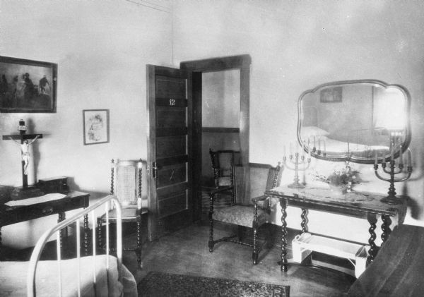 Interior of bedroom twelve at Saint Mary's Sanatorium, founded in 1880 by Bishop Salpointe. The view facing the door includes a bed, chairs, tables and a mirror. The table on the left holds a large crucifix.