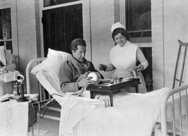 A male patient sits in bed on the porch and receives a meal from a nurse at Saint Mary's Sanatorium, founded in 1880 by Bishop Salpointe. A telephone sits on a table to the left.