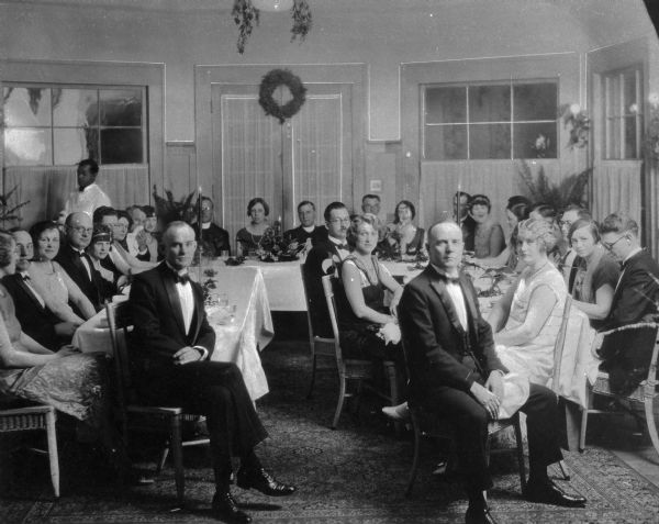 A group of patients in formal dress gathering around  tables for dinner at Saint Mary's Sanatorium, founded by Bishop Salpointe.  Windows and a set of doors can be seen in the background.