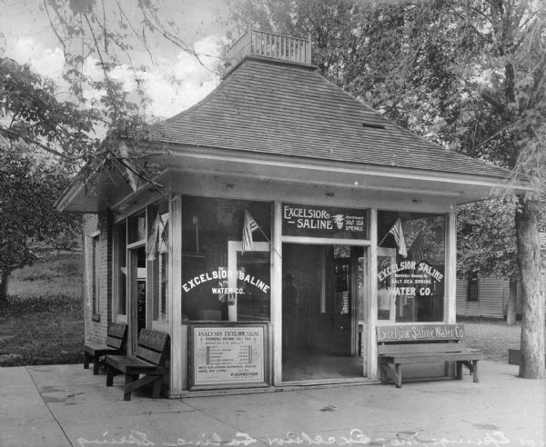 Exterior of an office at the Excelsior Saline Water Company.  Two United States flags mark the doorway, and benches line the exterior walls.  On the front of the office, a sign shows an analysis of the mineral water.  Above the doorway, the text reads, "Excelsior Saline - Formerly Known as Salt Sea Springs."