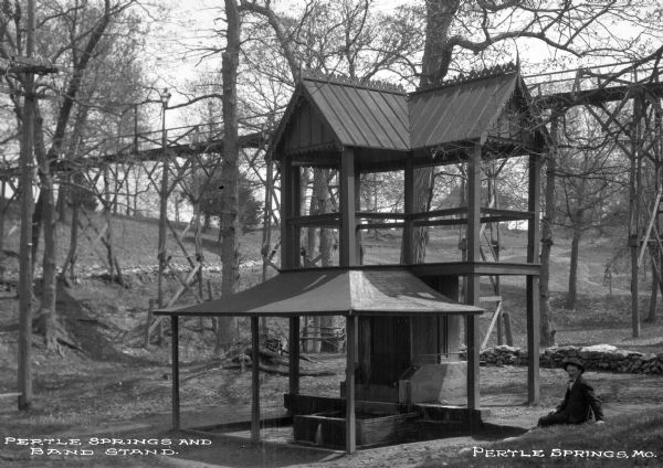 A view of a bandstand and Pertle Springs, an iron-rich spring whose name comes from William S. Purtle, the owner of the land in the early 1800's. A man sits to the right of the bandstand, and a bridge can be seen behind him. Caption reads: "Pertle Springs and Band Stand."