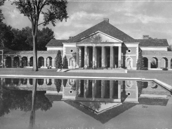 Front view of the large, monumental Administration Building with a reflecting pool. Also known as the Simon Burach Building after the famed hydrotherapist, it is located in the Saratoga Spa State Park and was built in the 1930's. A few people are gathered on the stairs. Flanking the building is an open-sided walkway with arches.