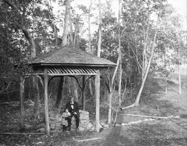 Wallace Bruce and a young girl rest at Alpine Park, near DeFuniak Springs. He is sitting on a well, and the girl is standing beside him under a covered shelter in a wooded area.