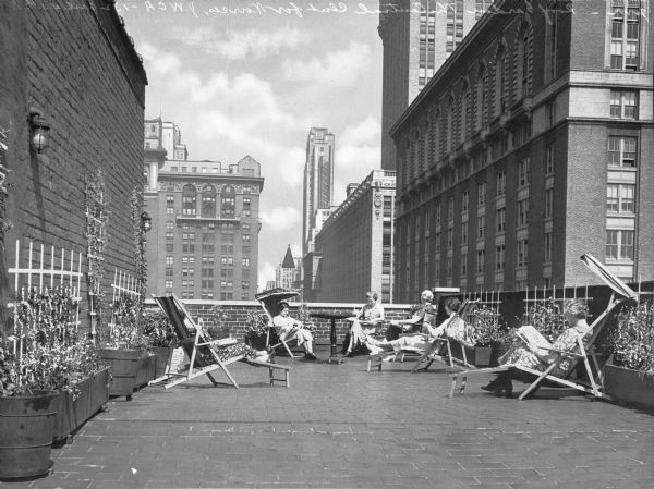 View of the roof garden at the Central Club for Nurses, completed in 1916 by architects Parish and Schroeder. The Central Club for Nurses was built for the Young Women's Christian Association, founded in 1855.  On the roof, nurses are seated, reading newspapers. Behind them are the tall buildings of the city.