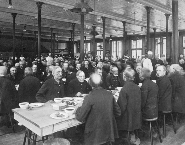 A large group of veterans eating in the dining hall at the National Military Home, which opened in 1867. Men in white hats and uniforms serve a meal to the veterans.