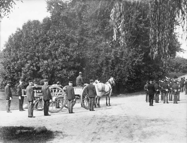 View of a funeral ceremony at the National Military Home, which opened in 1867. A group of veterans march before a horse-drawn vehicle pulling a coffin on a wagon.