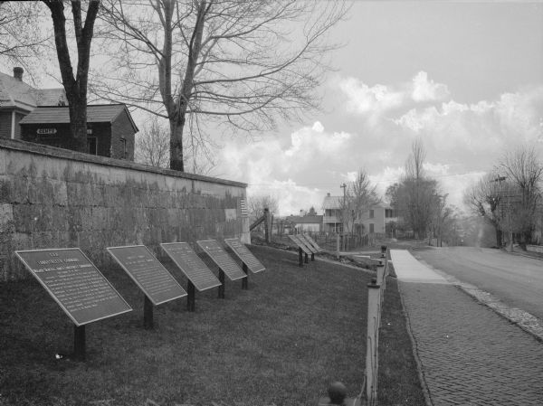 A view of eight commemorative tablets at the North Cemetery of Antietam Battlefield, the site of the bloodiest one-day battle in United States history in 1862. The tablets are memorials to Major General Longstreet's Command. A road curves to the right, and a stone wall and residential buildings can be seen in the the background.