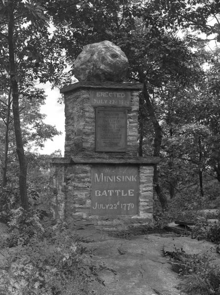 View of a monument to the Minisink Battle. The plaque reads, "Erected by the Historical Societies of the Minisink Country and of the State of New York on the sesquicentennial of the battle. 1929." The top portion of the monument reads, "Erected on July 22, 1874," and "Dedicated to the memory of the patriots of the Minisink region who died here July 22, 1779 in the defense of American Liberty." Atop the monument sits a large boulder.
