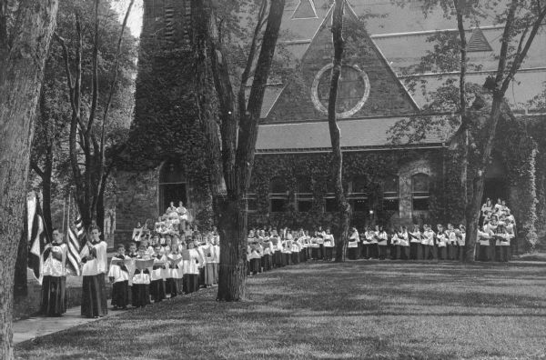 A children's choir stands outside Flemington Presbyterian Church, completed in 1883. The group of children process out of each doorway, holding song books and wearing choir uniforms. Two boys lead the choir, carrying flags.