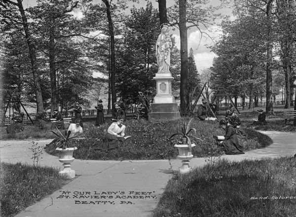 View of women reading at Saint Xavier Academy, established by the Sisters of Mercy in 1847. The women sit in the grass near a statue of Mary and Jesus. Caption reads: "'At Our Lady's Feet' St. Xavier's Academy, Beatty, PA."