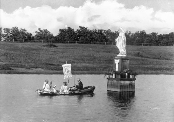 View of a statue of the Immaculate Conception. A man rows a boat toward the statue which stands in the water. A priest blesses the statue and two servers sit near him, one holding a banner.