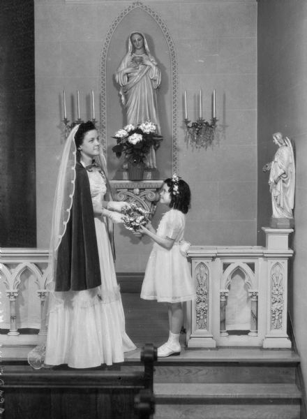 View of a young girl presenting a crown of flowers to a woman who will place it on the statue of Mary at Holy Family Academy, built in 1909. A pew is in the foreground, and behind the woman and the girl, a statue of Mary stands against the wall.