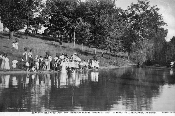View across water towards an outdoor baptism at McBrayer's Pond. A group of adults and children are gathered at the water's edge, some of them holding parasols. A man stands in the water next to a child in white clothing. Caption reads: "Baptizing at McBrayer's Pond at New Albany, Miss."