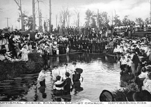 A large group of adults and children gather around a pond where a baptism is taking place, some standing on a bridge on the right. Several people stand in the water in the foreground, being baptized. Caption reads: "Baptismal Scene, Emanuel Baptist Church, Hattiesburg, Miss."