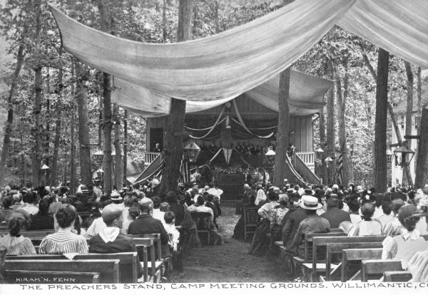 View toward a preacher's stand at the meeting grounds of the Willimantic Camp Meeting Association, established in 1860. A large group of people sit on outdoor benches facing the stand, listening during an outdoor meeting at the camp. Caption reads: "The Preachers Stand, Camp Meeting Grounds, Willimantic, Conn."