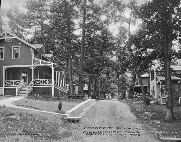 View of the meeting grounds of Willimantic Camp Meeting Association, established in 1860. Two women walk toward the foreground on Prospect Avenue, a dirt road. A sign over the stairs of the building on the left reads, "Woodland Nook." Caption reads: "Prospect Avenue. Willimantic Camp Meeting Grounds."