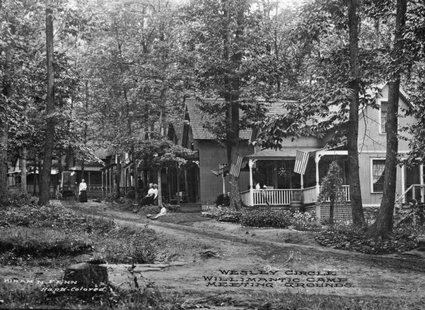 View of the camp meeting grounds of the Willimantic Camp Meeting Association, established in 1860. Women and a child gather near a row of homes on Wesley Circle. Caption reads: "Wesley Circle Willimantic Camp Meeting Ground."