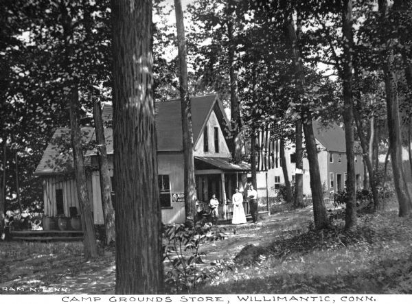 View of the campgrounds store of the Willimantic Camp Meeting Association, established in 1860. A man, woman, and child stand at the porch of the store, under a United States flag. Caption reads: "Camp Grounds Store, Willimantic, Conn."