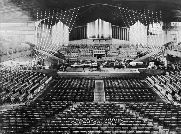 View from balcony of Ocean Grove Auditorium, built in 1894. The vast seating provided space for the Ocean Grove Camp Meeting Association. A platform stands in the center of the auditorium, and behind it is a Hope-Jones pipe organ which was installed in 1908. Caption reads: "The Auditorium. Ocean Grove, N.J."