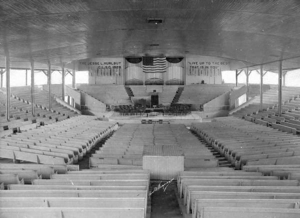 Interior of an amphitheatre at Lake Chautauqua, founded in 1874. Surrounding a platform, man wooden benches are seen. Above the center platform, a flag hangs in front of organ pipes. On one side of the flag, the wall reads, "The Jesse L. Hurlbut C.L.S.C. 1928," and on the other, "Live Up to the Best that is in You."