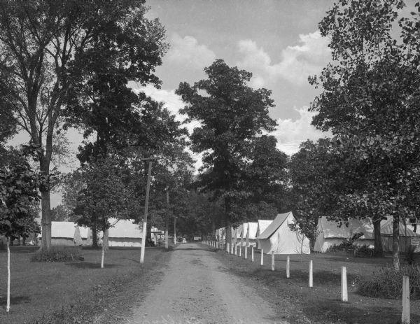 View of a row of tents along a dirt road at the Chautauqua Grounds.  Trees and white posts line the road.