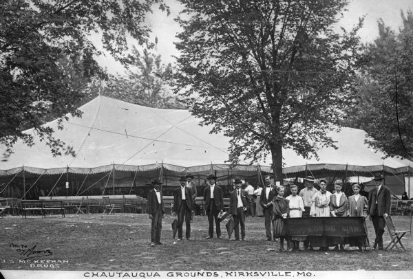 A group of men and girls stand in front of a large tent at Chautauqua Grounds, an establishment founded in 1874. The girls hold a sign that reads, "J.F. Egkert Leading Dry Goods Man." Caption reads: "Chautauqua Grounds, Kirksville, MO."