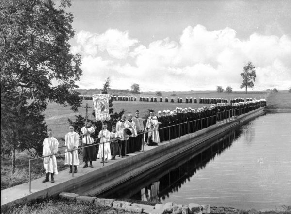 View of a procession to Immaculate Conception Lake at Nazareth Academy, established in 1814. Priests and servers lead the procession, and women in black follow, walking along Immaculate Conception Lake.