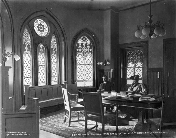 View of the reading room of the First Church of Christ Scientist, built in 1903. A man and a woman are sitting at a table surrounded by chairs and wooden benches. Stained glass windows line the walls. Caption reads: "Reading Room, First Church of Christ Scientist."