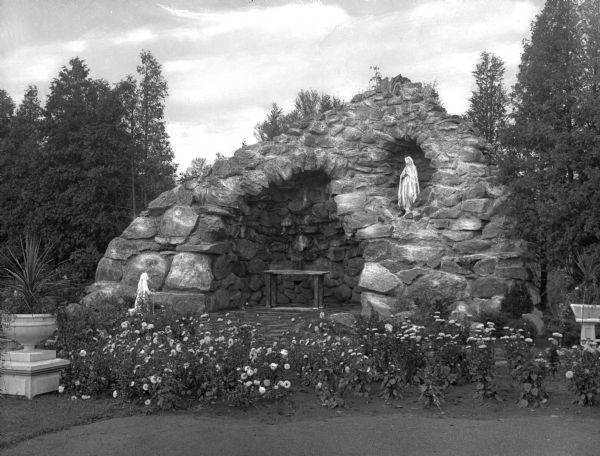 View of the grotto at the Putnam Catholic Academy. Flowers surround the grotto with a statue and an altar inside.