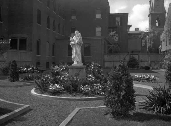 View of Saint Joseph's Garden and statue at Saint Joseph's Guest House. Flowers and plants surround the statue, and church buildings can be seen in the background.