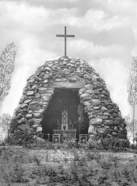 Front view of a stone grotto at Saint Louis Academy, founded in 1818. Two kneelers stand before an altar, and a statue can be seen to the right. Atop the grotto stands a wooden cross.